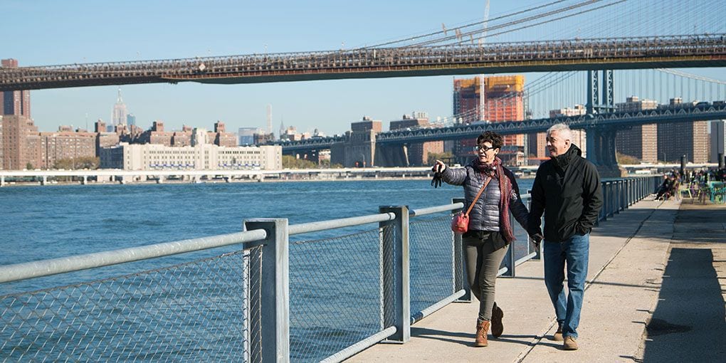 An older couple walking together along a riverside path on a clear day.