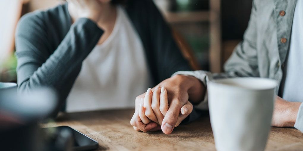Two people sitting at a kitchen table, one holding the other's hand.